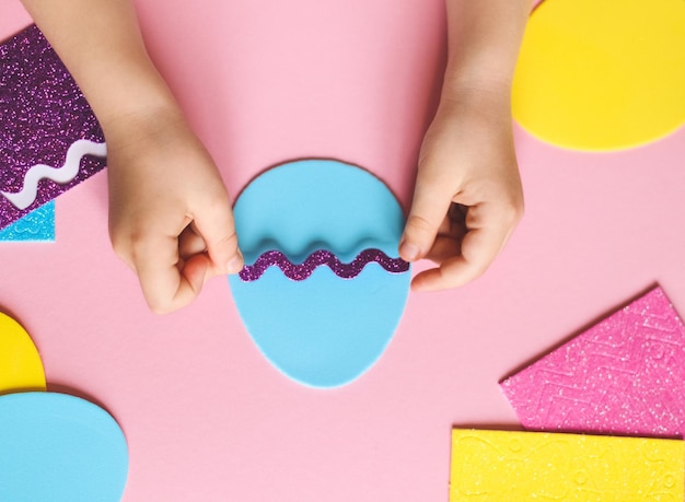 The hands of a little caucasian girl glue a lilac zigzag sticker on a felt egg sitting at a table