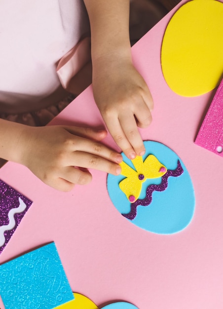 The hands of a little caucasian girl stick a yellow bow sticker on a felt egg while sitting a table