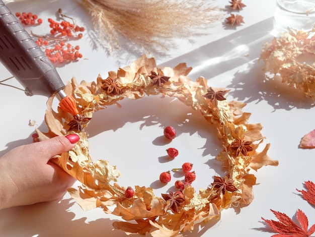 Hands making dried floral wreath from dry Autumn leaves and Fall berries Hands with manicured nails fix decorations with glue gun