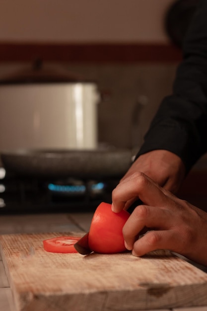 Hands of the male chef slicing a fresh red tomato on the rustic wooden board with a knife in the kitchen