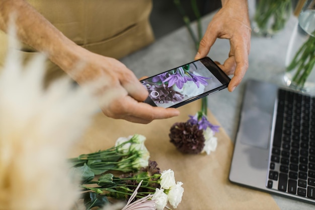 Photo hands of male florist holding smartphone taking picture of flower on counter