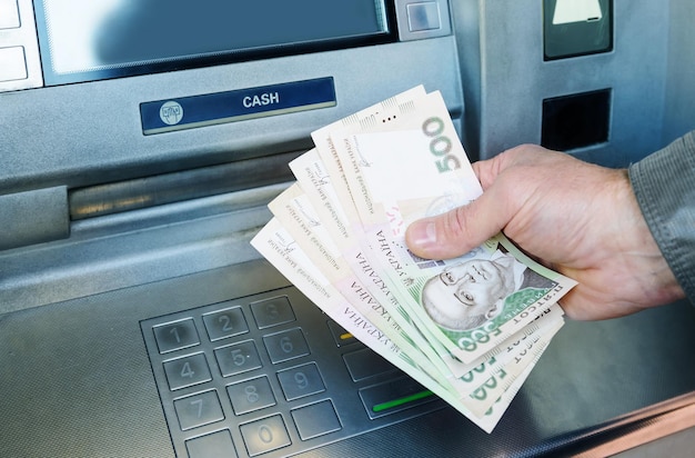 Hands of a man are holding banknotes of the Ukrainian hryvnia on the background of ATM.