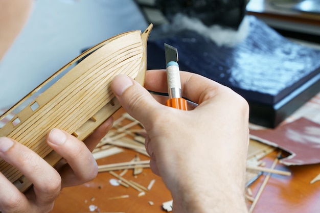 Hands man cutting out details for ship model with clerical knife from plywood Process of building toy ship handicraft