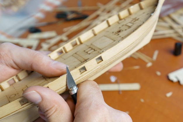 Hands man cutting out details for ship model with clerical knife from plywood Process of building toy ship handicraft