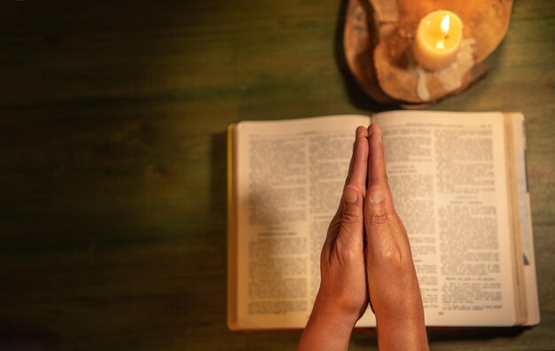 Hands of man in prayer posture on a bible illuminated with a wooden candle