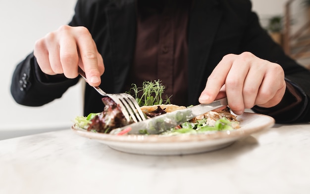 Hands of a man with a fork, a knife and a plate with a salad close-up. A man eats a salad at a restaurant table. Background. Copyspace