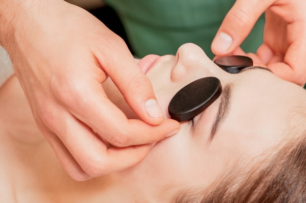 Hands of massage therapist puts massage stones on eyes of young woman during relaxing massage.