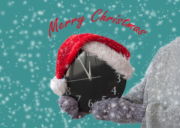 Hands in mittens to hold the watch which is wearing a Santa Claus hat around the snow postcard merry Christmas