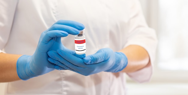 The hands of a nurse in blue gloves and a white coat are carefully holding a bottle of coronavirus vaccine. Close up. Wide banner