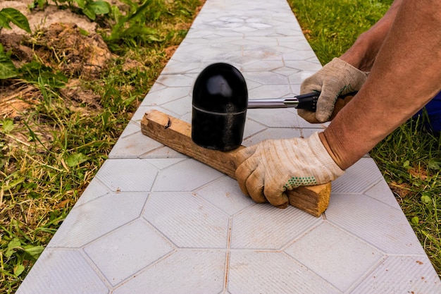 The hands of a professional worker hit the slab with a special rubber hammer in the process of laying a garden path made of stone tiles