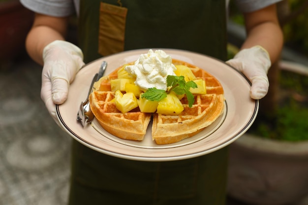 Hands in rubber gloves holding Belgium waffles with pineapple and ice cream on a plate