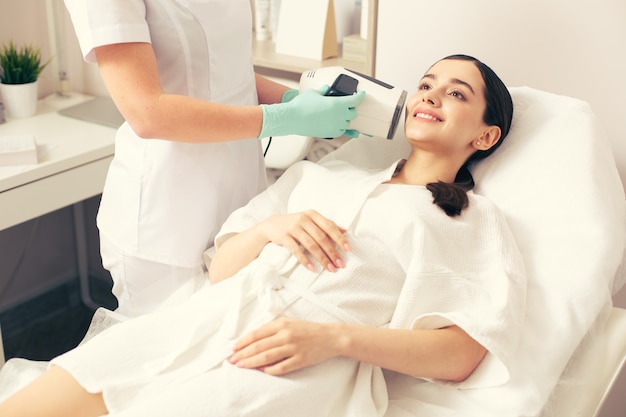 Hands in rubber gloves holding a modern skin analysis camera near the face of a young smiling woman