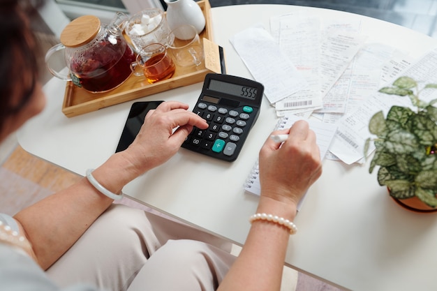 Hands of senior woman using calculator to check bills and sum up monthly expenses