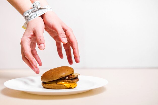 Hands take a burger, centimeter ties hands, violation of diet, hands reaching for a burger which lies on a plate on a white background