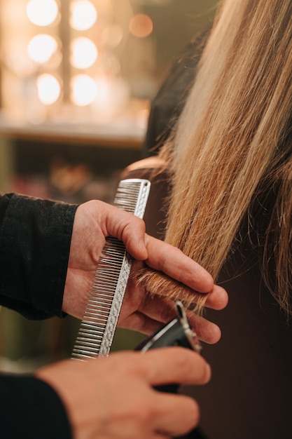 Hands of an unrecognizable person doing a haircut with a razor to an unrecognizable girl with long blond hair. Vertical