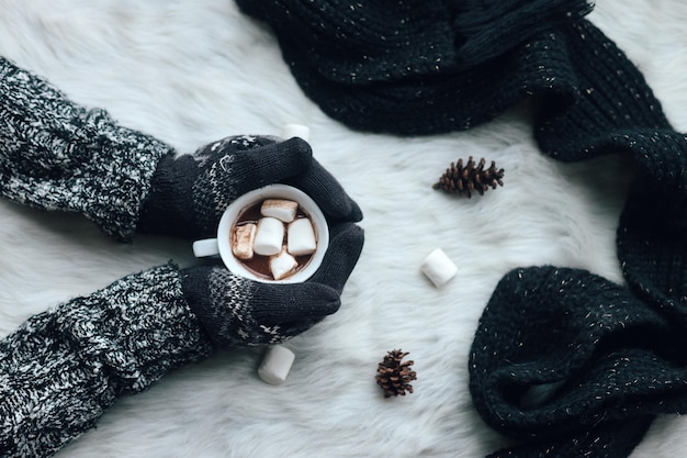 Hands using warm winter clothes and gloves hold a cup of hot chocolate on white fur rug