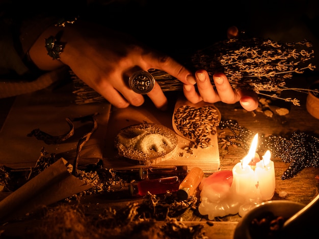 In the hands of the witches bunch of dry herbs for divination. The light from the candles on the old magic table. Attributes of occultism and magic.