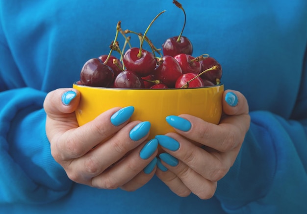 Hands with a bright blue manicure hold a yellow bowl with cherries