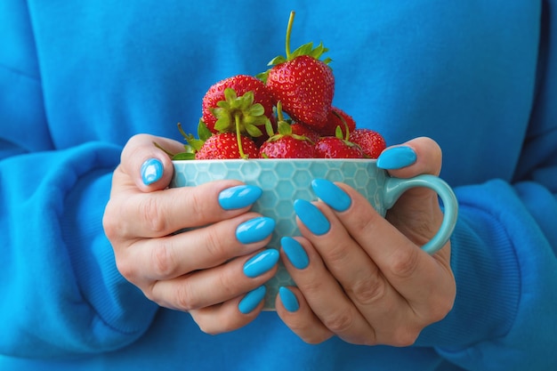 Hands with a bright blue manicure hold a yellow bowl with strawberries