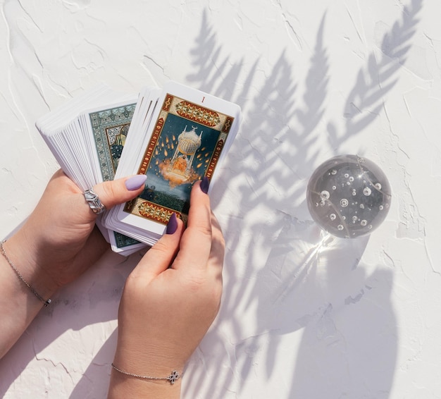 Photo hands with purple nails and rings hold deck of tarot cards on white surface. crystal ball and fern leaf shadow, top view