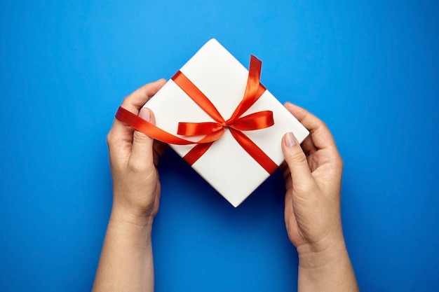 Hands with white gift box tied with red ribbon on blue