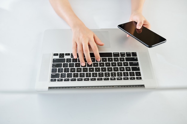 Hands of woman checking smartphone and working on laptop, view from above