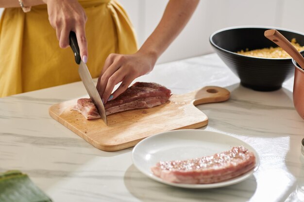 Hands of woman cutting seasoned pork on kitchen table