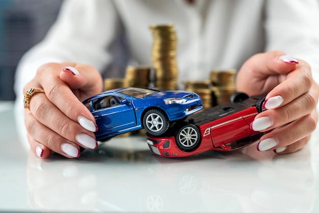 Hands of a woman holding two car models simulating an accident near a mountain of coins Car insurance Car accident driving safety