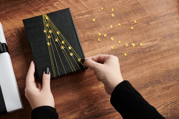 Hands of woman sticking sparkling stars on giftbox wrapped in black paper