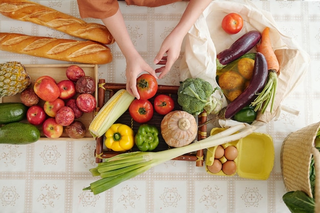 Hands of woman unpacking recyclable bags of fruits and vegetables at kitchen table after coming home from market, view from the top