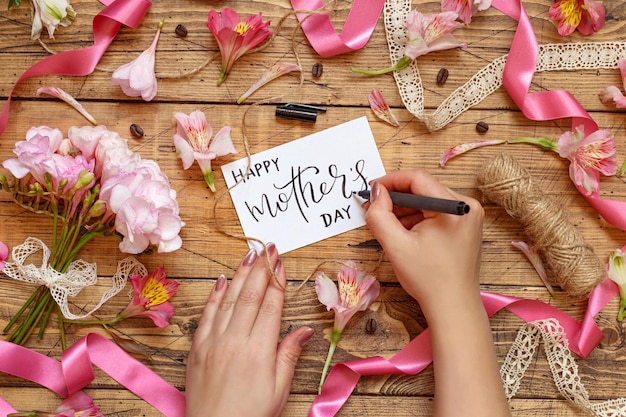 Photo hands writing happy mothers day card on a wooden table between pink flowers top view