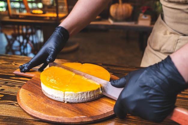 Hands of young chef baker cutting slice of mango cheesecake