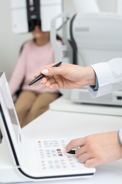 Hands of young contemporary female ophthalmologist with pen pointing at display while examining eyesight of elementary schoolgirl in clinics