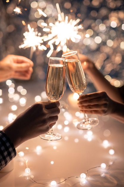 Hands of young couple clinking with flutes of champagne on space of two humans holding sparkling bengal lights