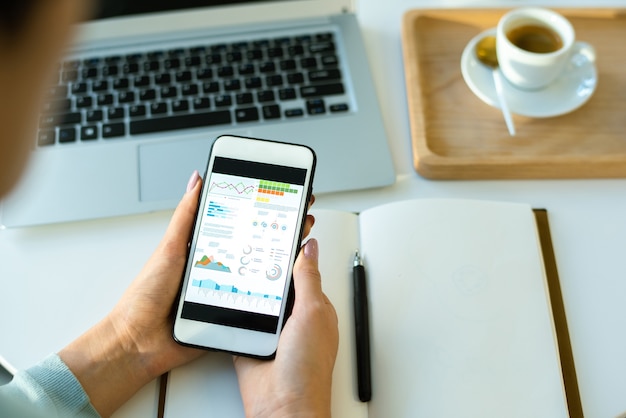 Hands of young economist over open notebook scrolling through financial data in smartphone while working by table in cafe