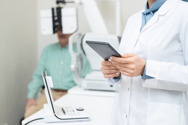 Hands of young female ophthalmologist in whitecoat using digital tablet on background of senior male patient having his eyesight checked up