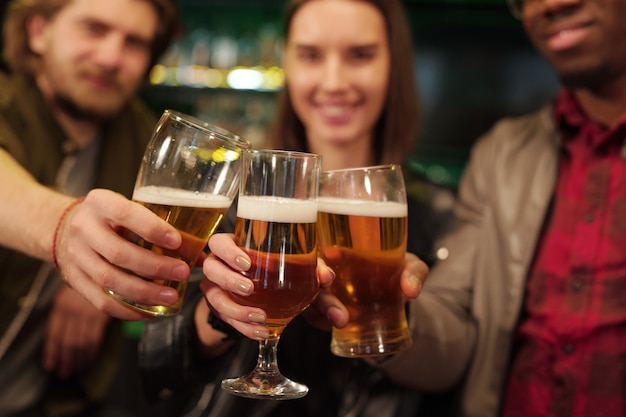 Hands of young intercultural football fans clinking with glasses of beer in front of camera while celebrating victory of their favorite football team