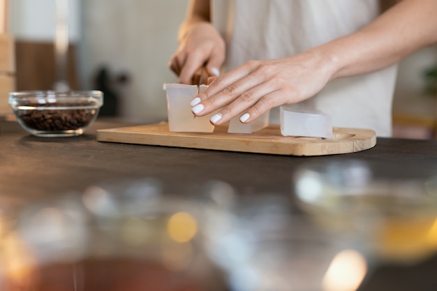 Hands of young woman with knife cutting piece of hard soap mass on wooden board while making natural cosmetic products at home