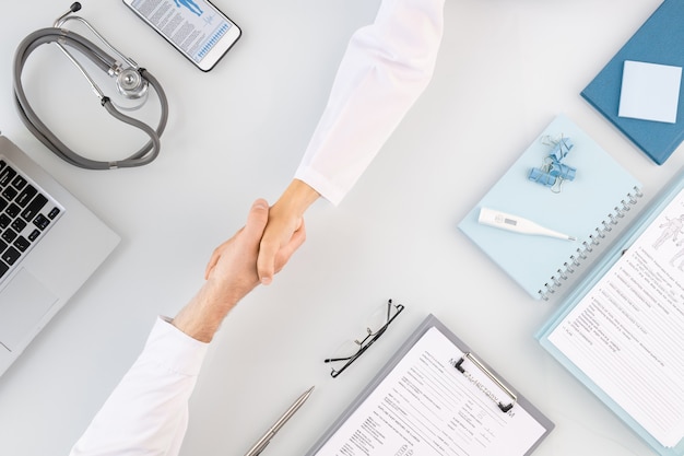 Photo handshake of two young successful clinicians in whitecoats greeting one another over workplace with documents and other medical supplies