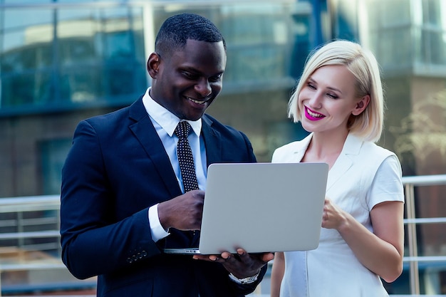 Handsome African American businessman in a stylish black suit working laptop with attractive blonde woman business lady on the street background office skyscraper successful deal idea