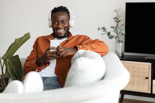 Handsome african american man in headphones listening to music on cellphone sitting on couch and smiling at camera