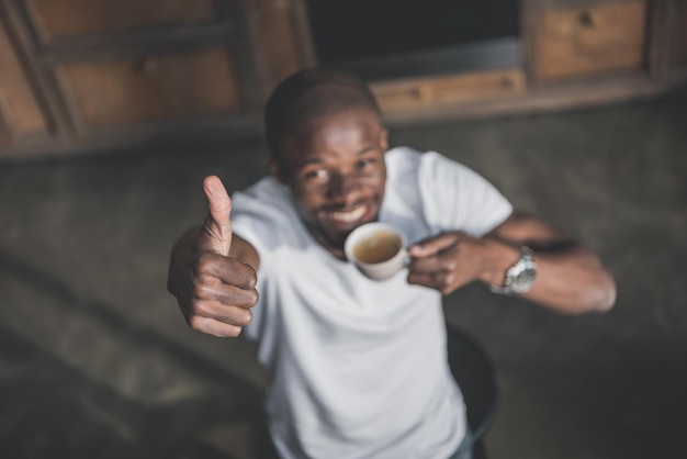 Handsome african american man showing thumb up and having his morning coffee at home