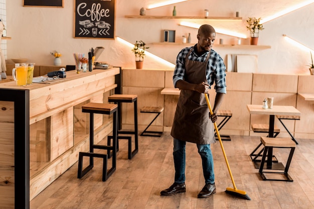 Handsome african american worker sweeping with broom in coffee shop