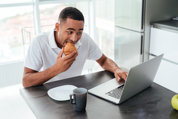 Handsome african american young man having breakfast and using laptop on the kitchen