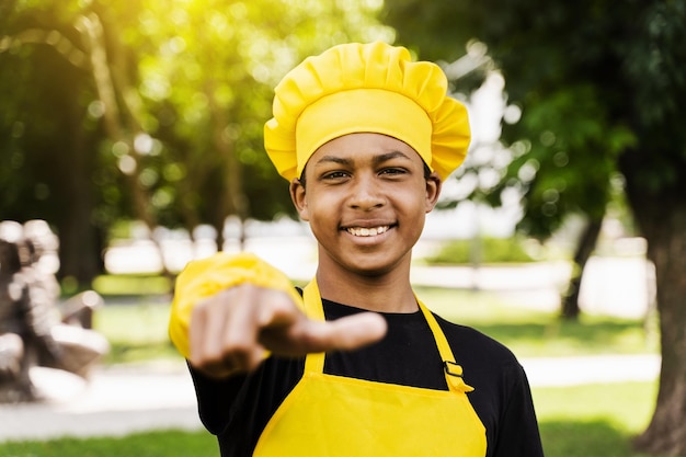 Handsome african teenager cook points to you Black child cook in chefs hat and yellow apron uniform smiling and pointing to you outdoor Creative advertising for cafe or restaurant