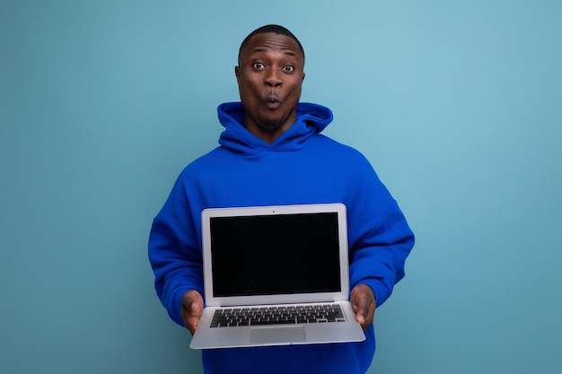 Handsome african young consultant in hoodie demonstrating open laptop mockup against background with