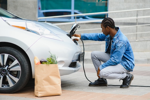 Handsome afro american guy sitting near his new modern electric car and holding plug of the charger