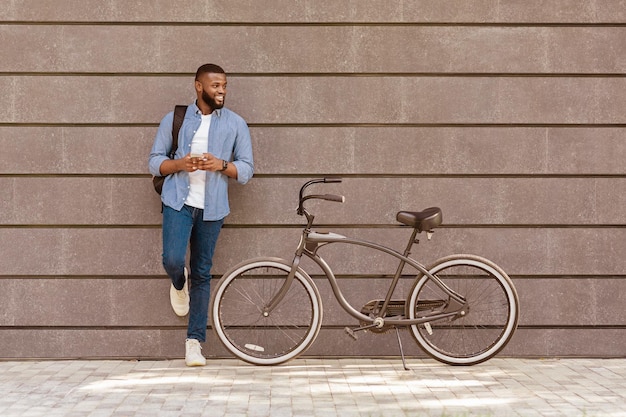 Photo handsome afro guy with backgpack and smartphone standing near his bike leaning on gray urban wall