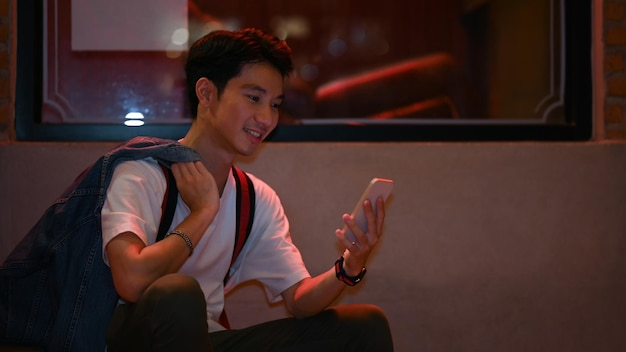 Handsome Asian male sitting near showcase in night city street and using mobile phone