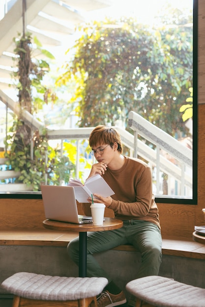 Handsome asian man using laptop sitting on wooden table in coffee shop with a cup of coffee